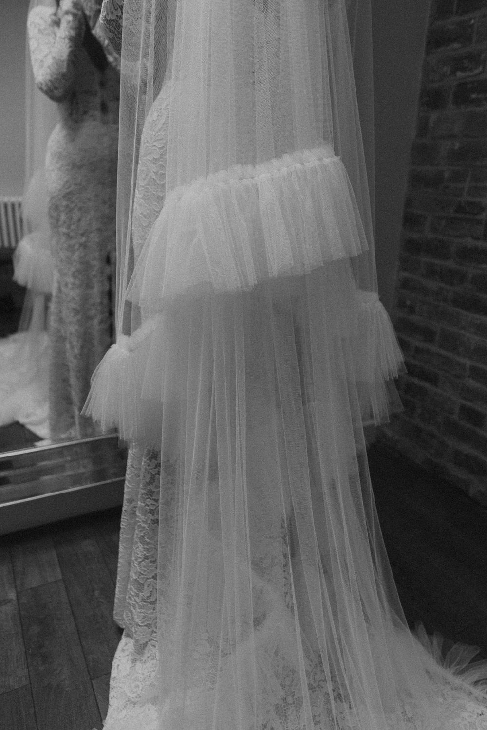 Close-up view of a flowing white wedding dress veil with delicate lace details against a brick wall background.