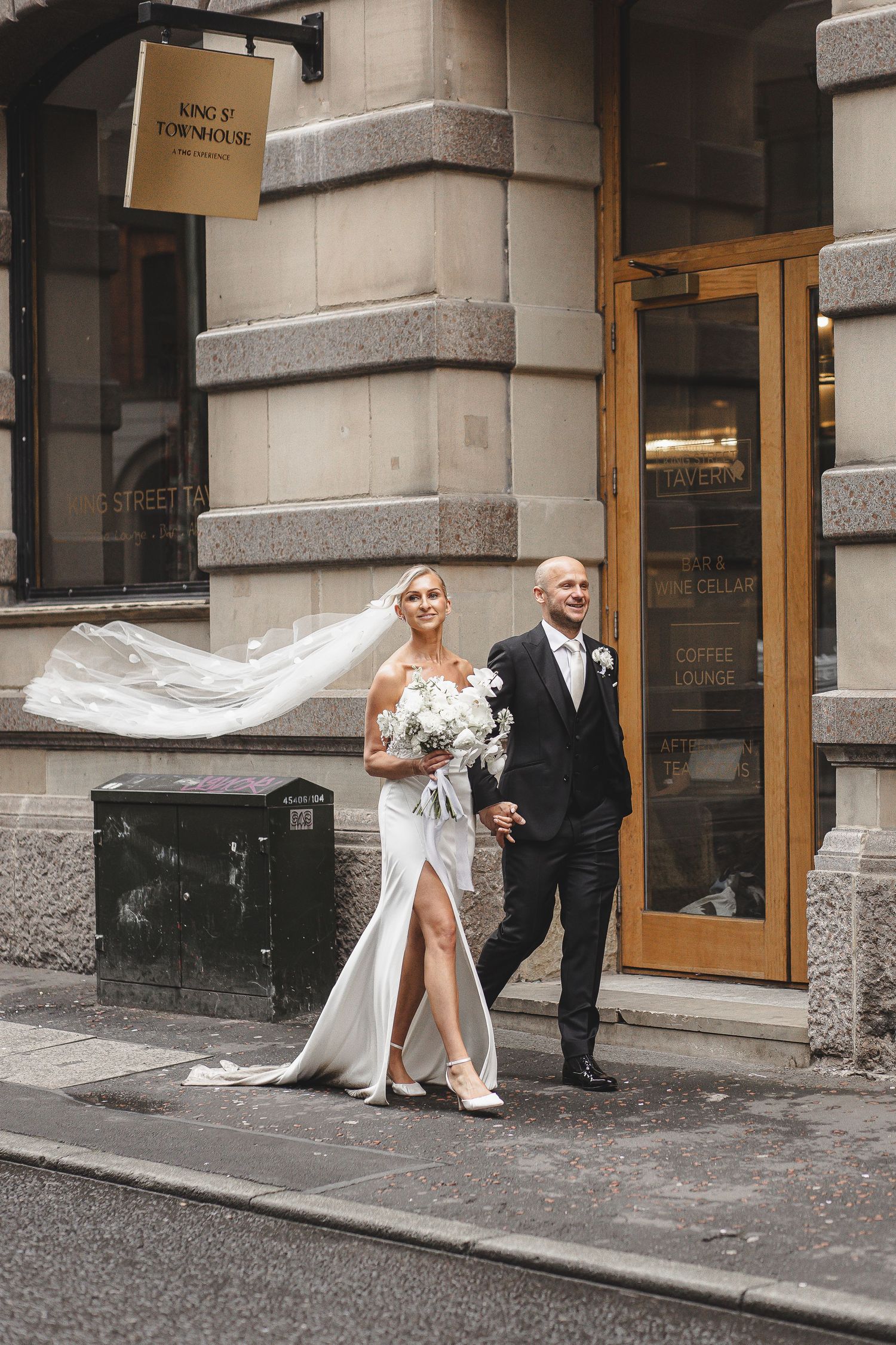 A couple in formal wedding attire stands outside an elegant stone building entrance.