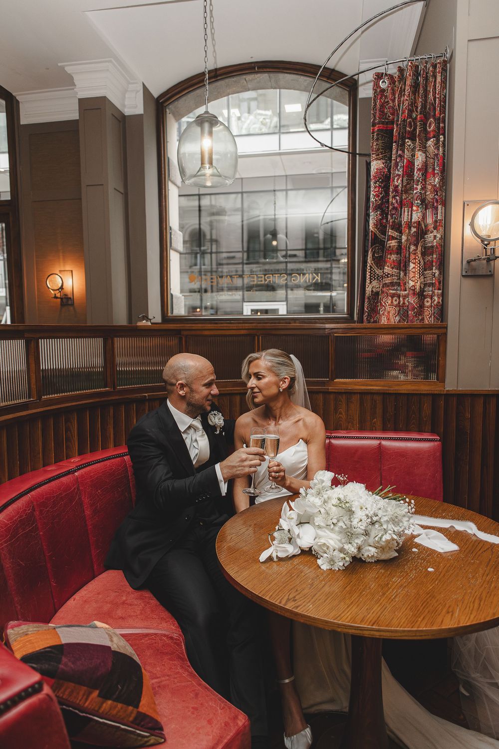 A couple shares an intimate moment at a restaurant booth with red leather seating and wooden tables.