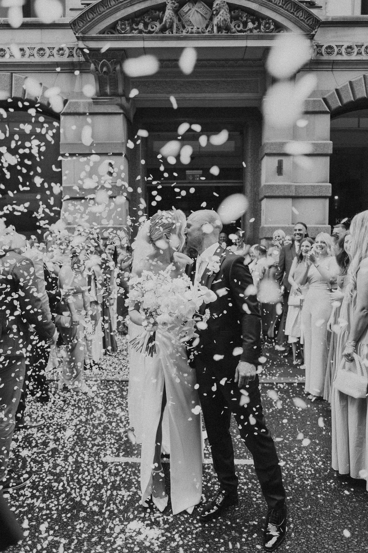 A vintage black and white wedding photograph showing a couple surrounded by confetti and guests outside an ornate building.