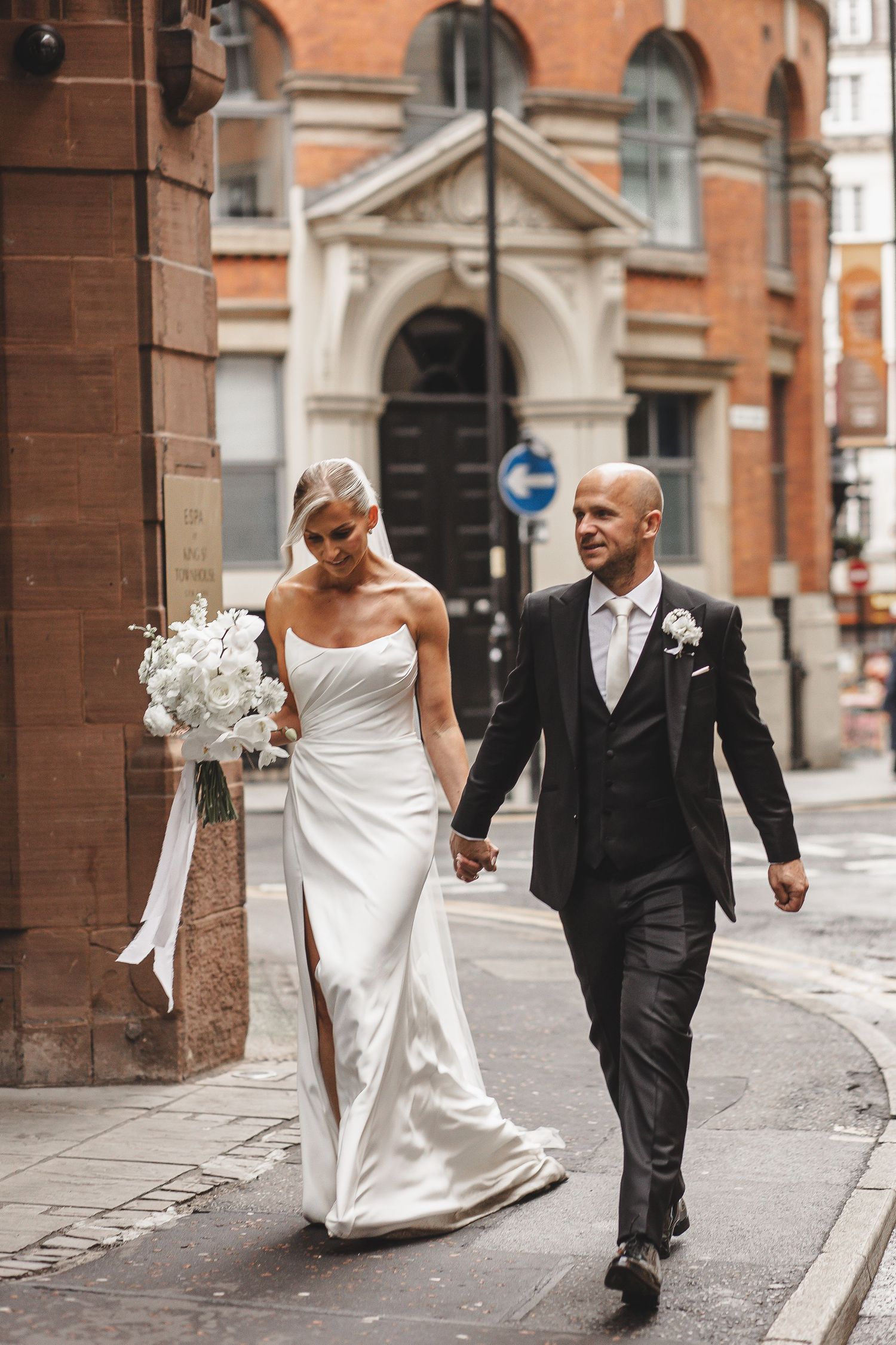 A bride and groom walk together on a cobblestone street against a backdrop of historic brick buildings.