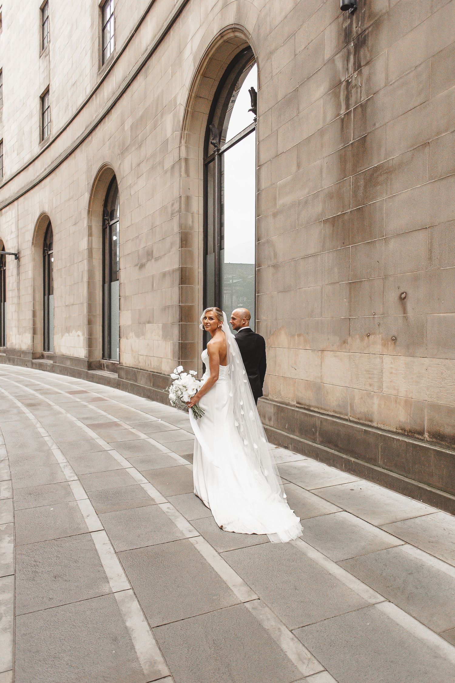 A bride in a white wedding dress walks gracefully through a stone archway corridor in an elegant architectural setting.