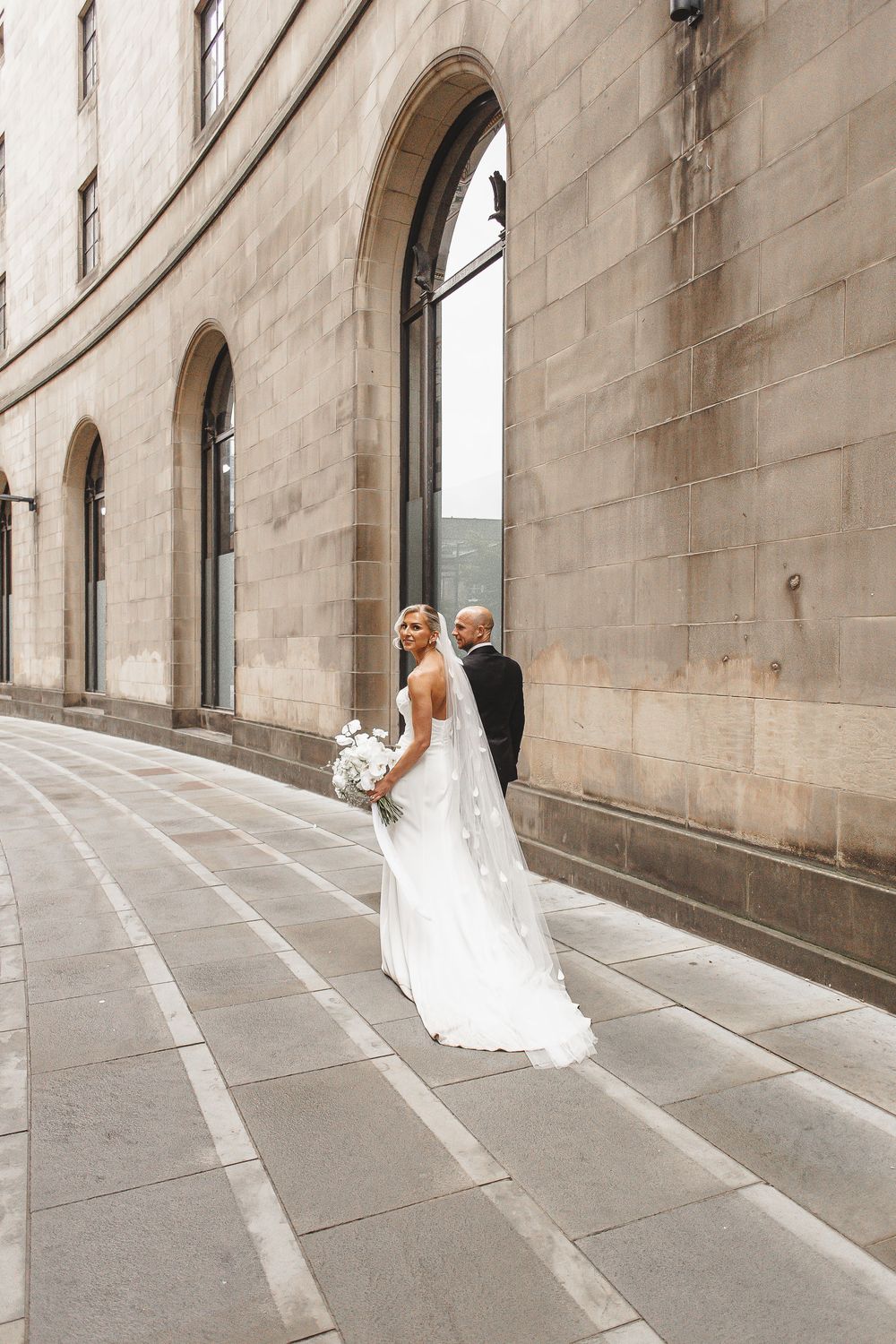 A bride in a white wedding dress walks gracefully through a stone archway corridor in an elegant architectural setting.