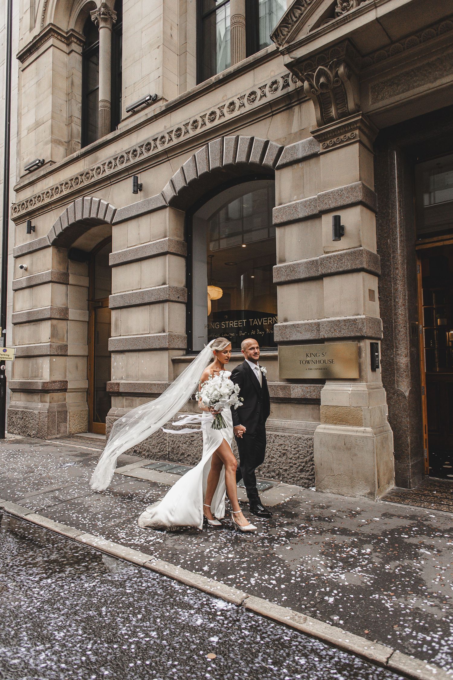 Newlywed couple poses playfully with flowing veil against historic stone building facade.