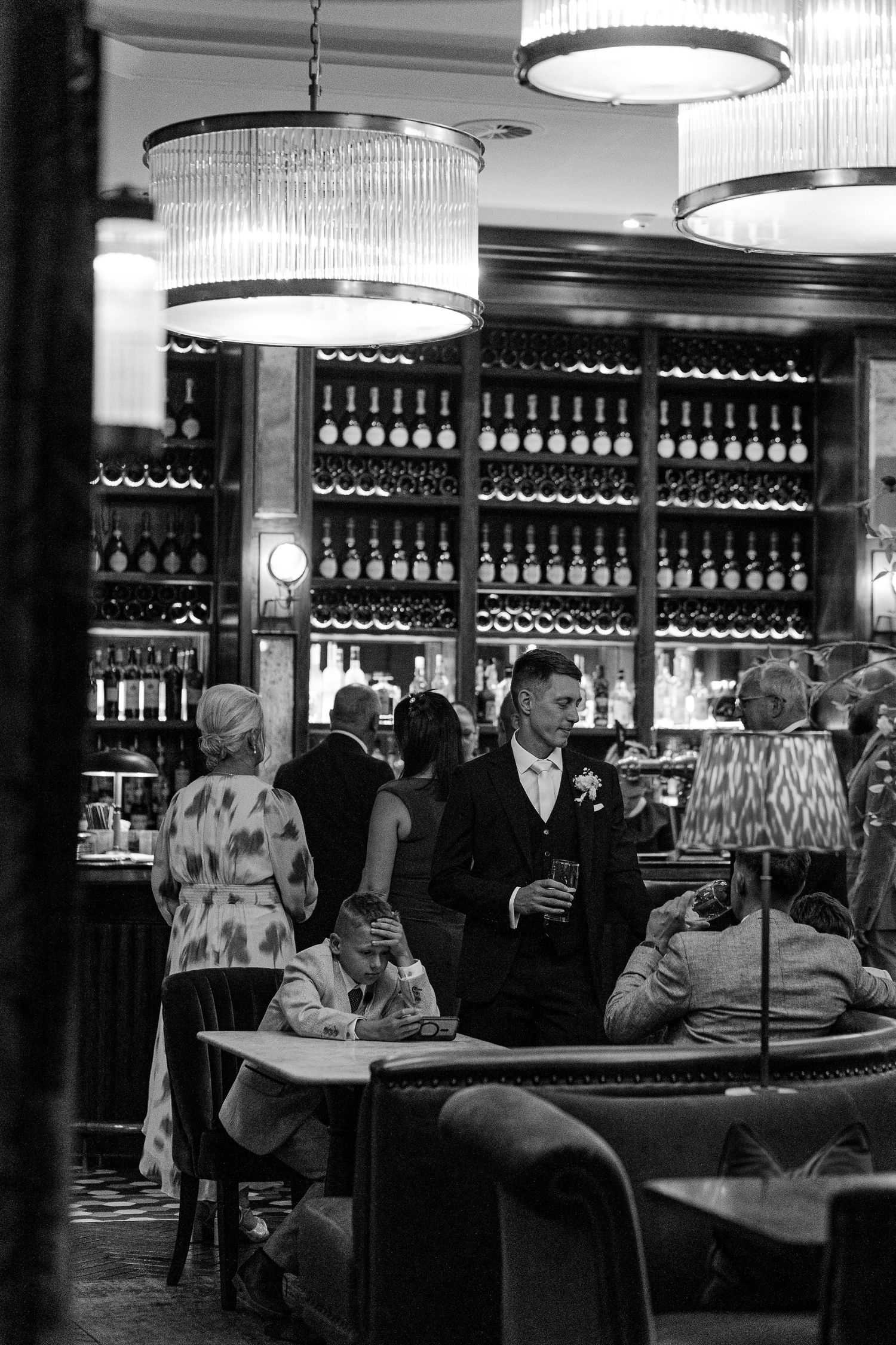 Black and white photos of wedding guests mingling at an elegant bar with wine bottles displayed on shelves.