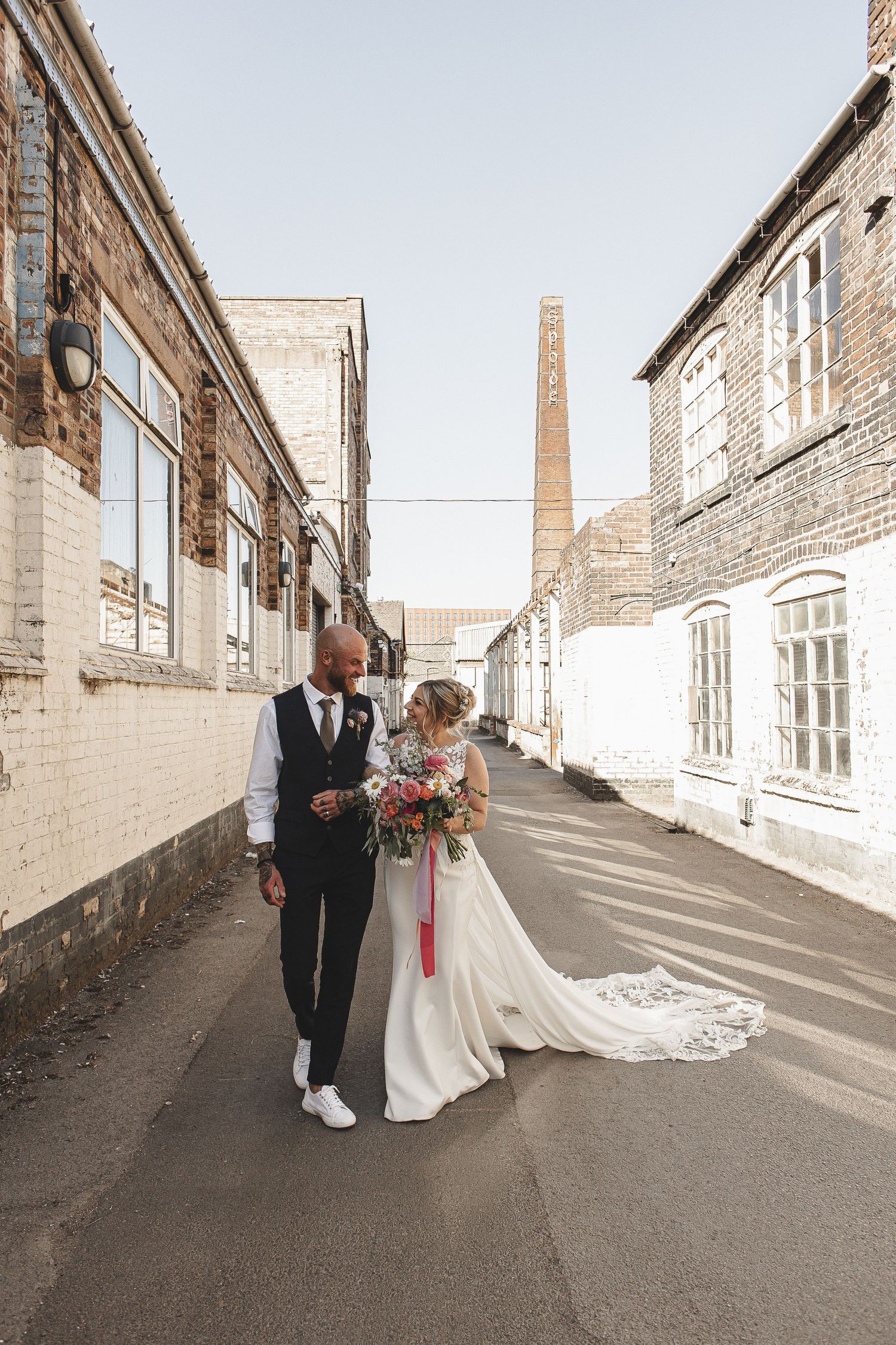 A couple walks together down an industrial alley with brick walls and windows on a sunny day.