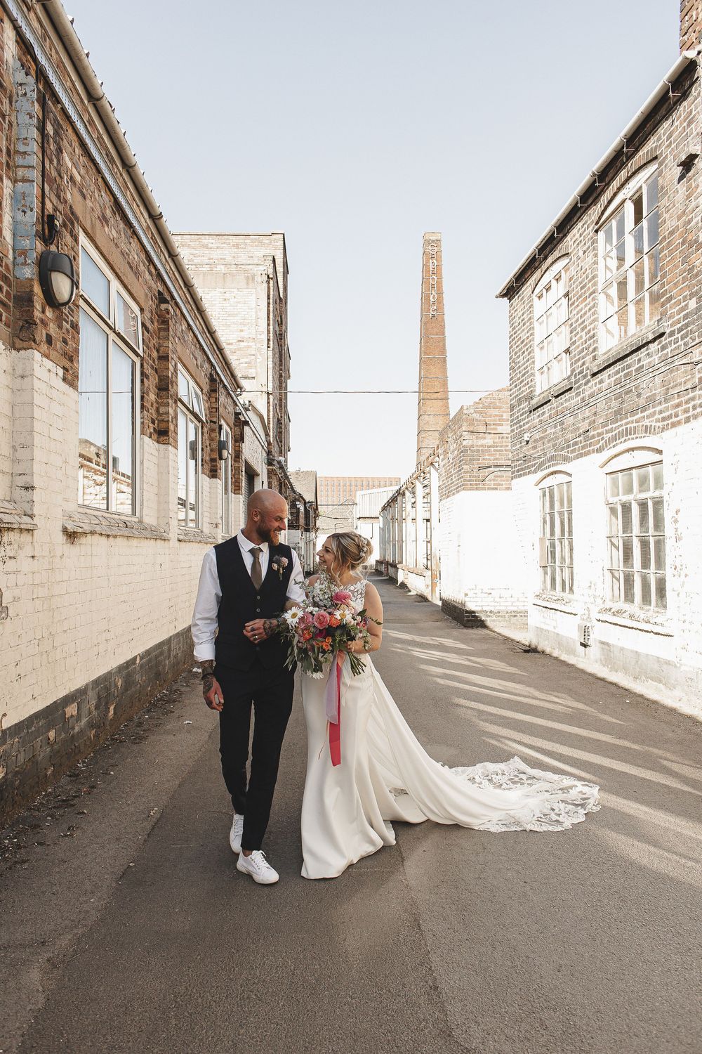 A couple walks together down an industrial alley with brick walls and windows on a sunny day.