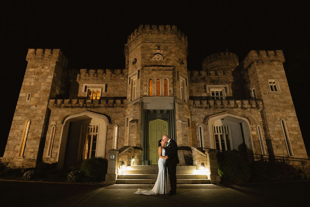 Bride and groom embracing at night in front of an illuminated stone castle wedding venue.