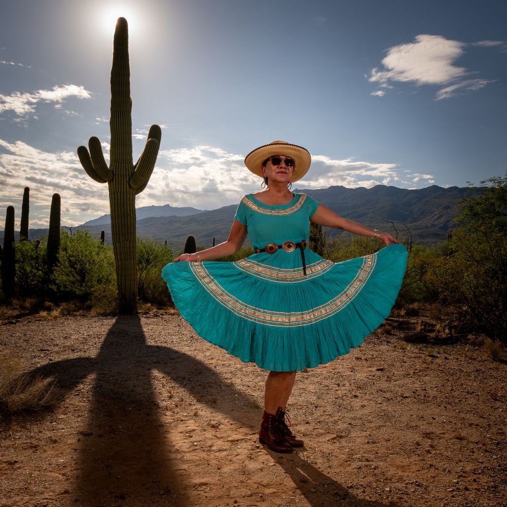 Person in turquoise dress and sun hat poses with saguaro cactus in desert landscape.
