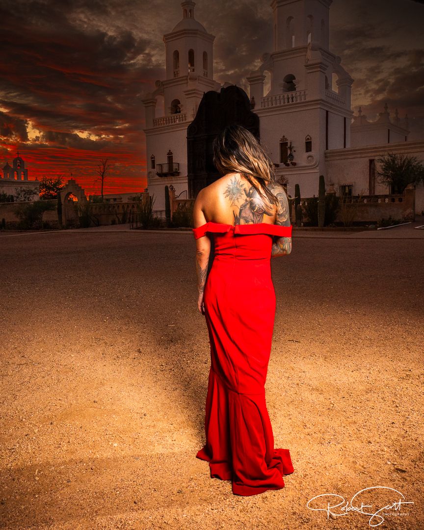 Woman in red dress facing white church building during dramatic sunset.