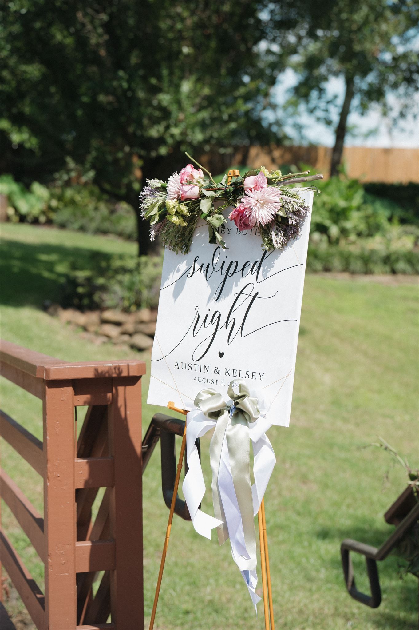 White wedding welcome sign decorated with pink and white flowers and ribbons stands on a wooden post in a garden setting.