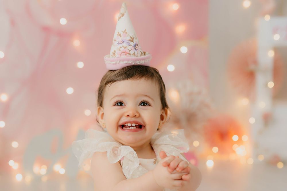 Fotografía de la fiesta del primer cumpleaños de una niña sonriendo con un cono floral  y decoración en tonos rosas