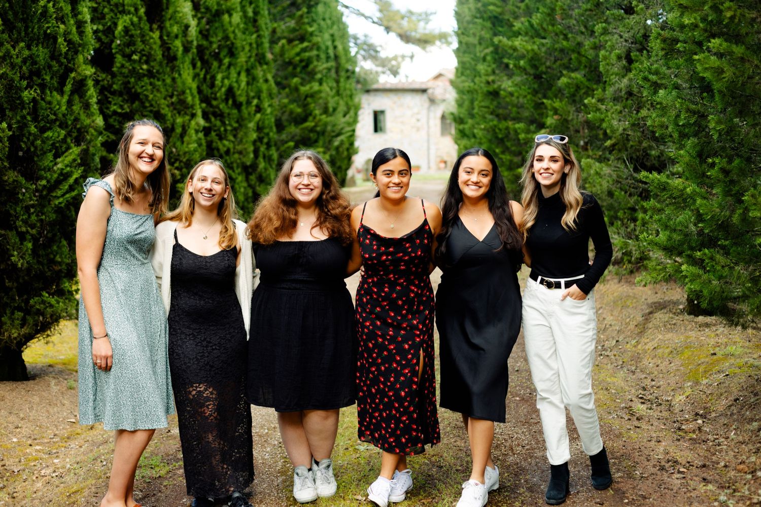 A group of friends in dresses pose together on a path lined with evergreen trees.