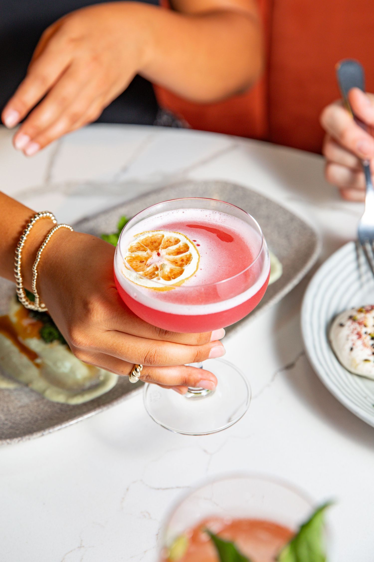 Pink cocktail garnished with citrus in a coupe glass held by someone wearing gold bracelets.