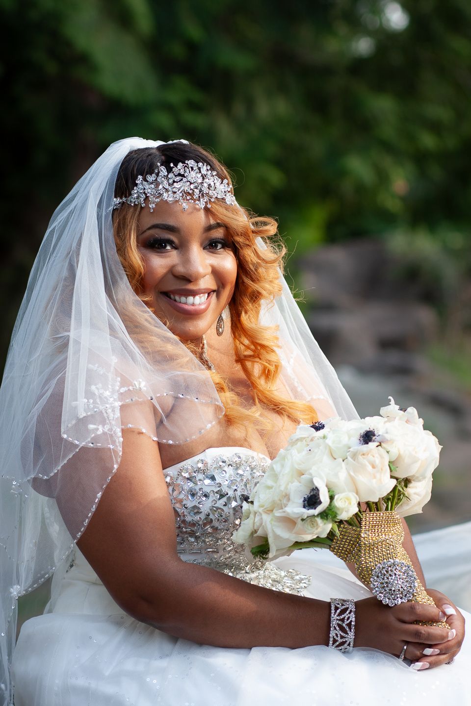 A bride in a white wedding dress and crystal tiara holds a bouquet of white flowers against a natural outdoor backdrop.