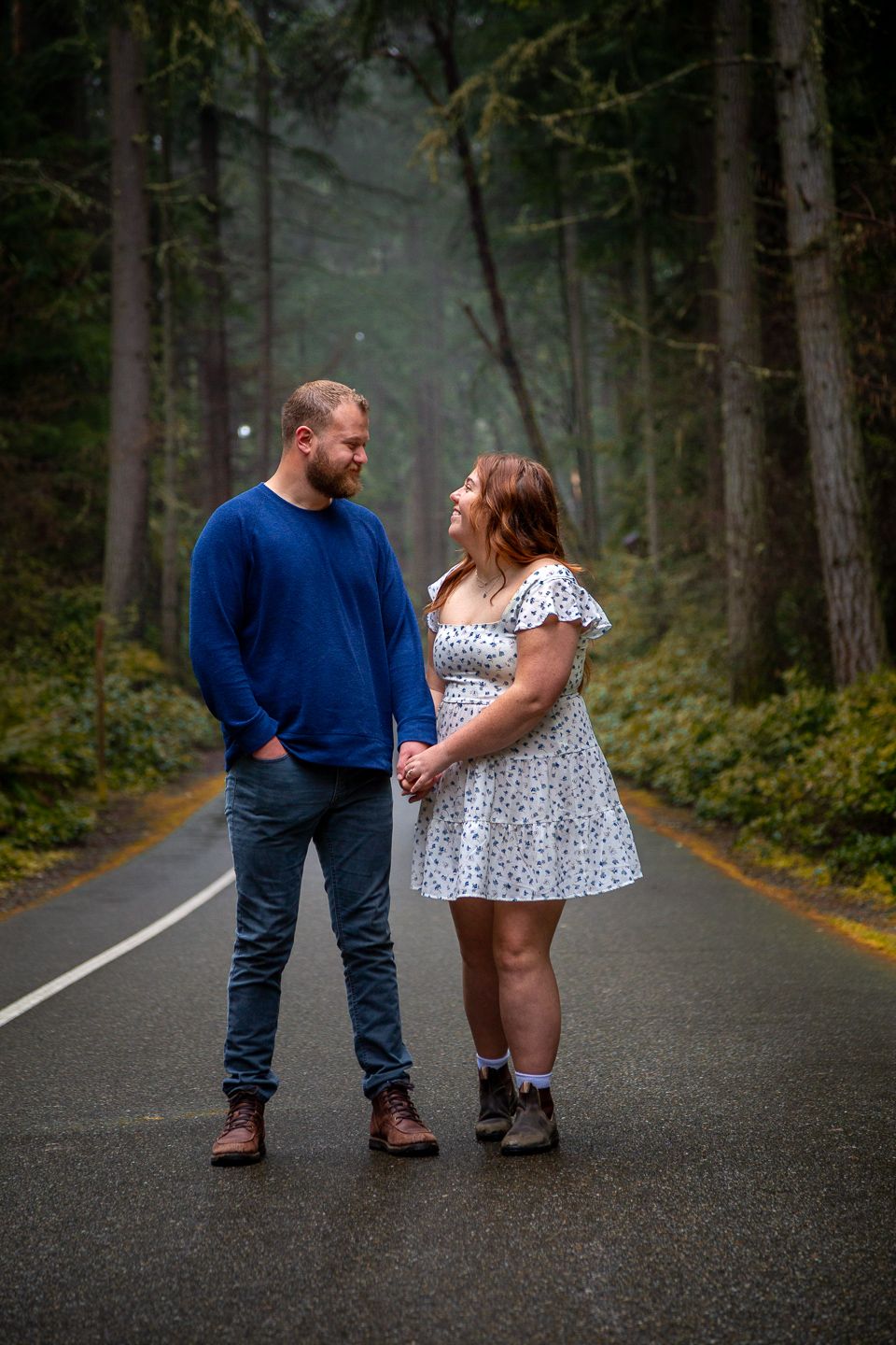Couple in casual attire standing together on a misty forest road surrounded by tall evergreen trees.