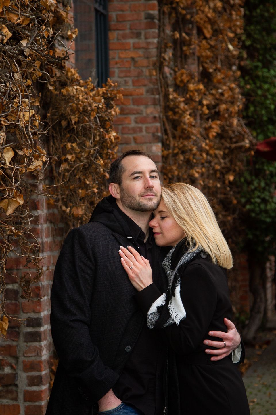 Couple in black winter coats embracing against rustic brick wall with dried vine backdrop.