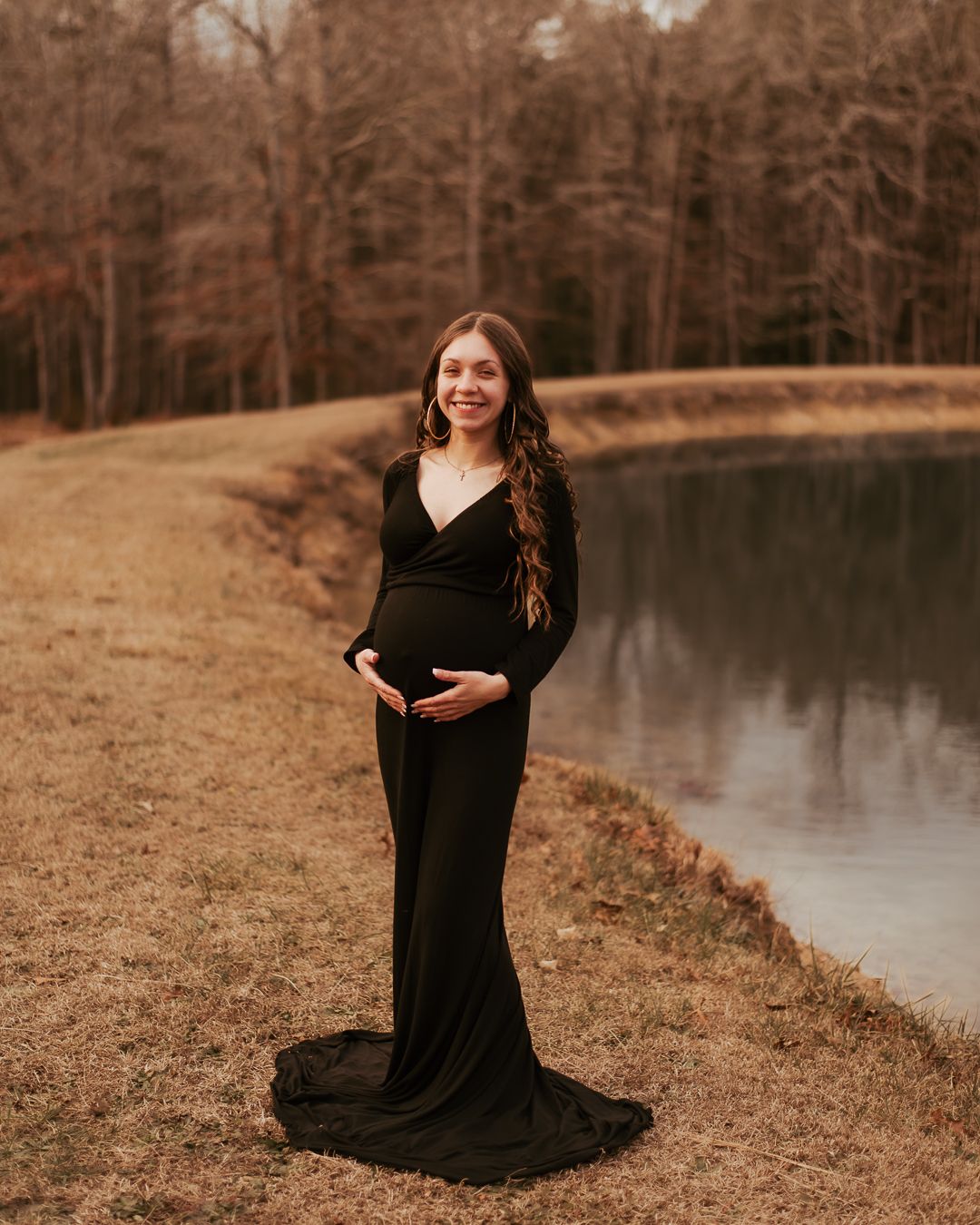 Maternity portrait by lake with woman in flowing black dress at sunset.