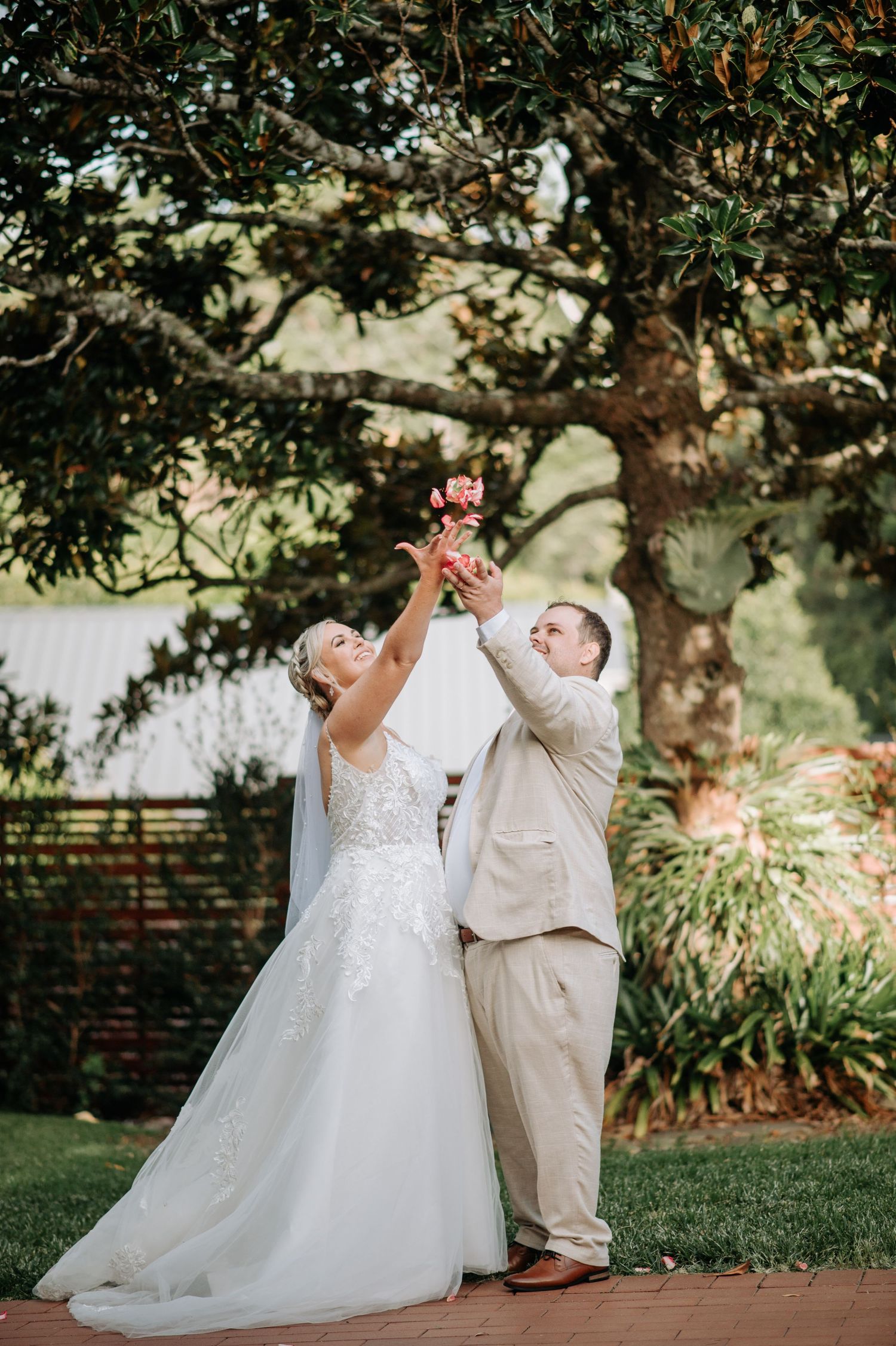 Newlyweds joyfully raise their hands together in celebration under a tree.