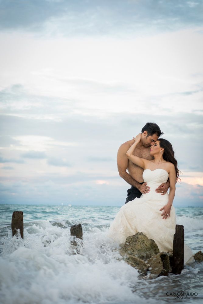 Dramatic wedding photo with waves crashing against rocks as couple embraces.