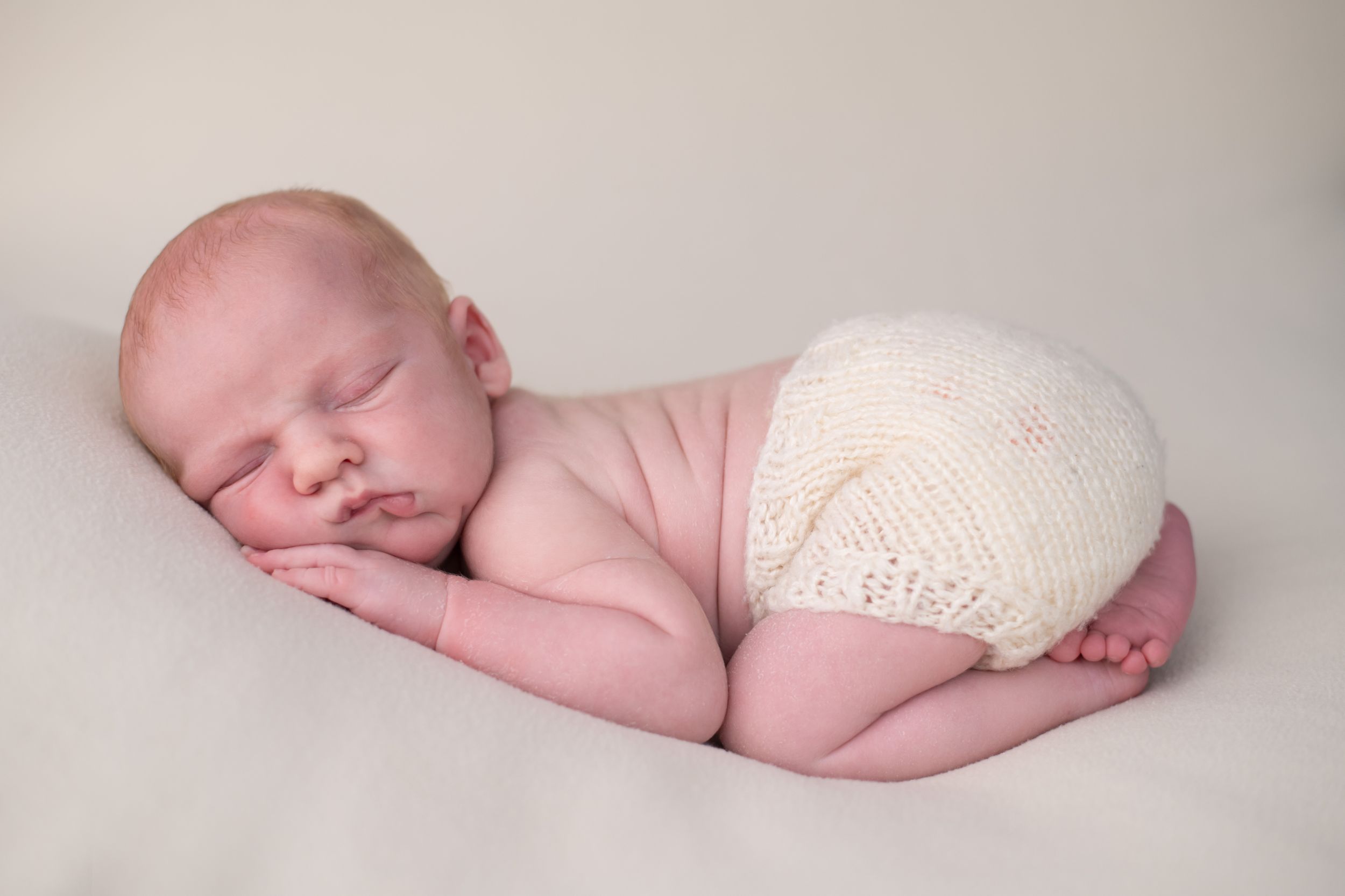 Sleeping newborn baby wrapped in soft cream-colored blanket lying on neutral backdrop in professional studio setting.