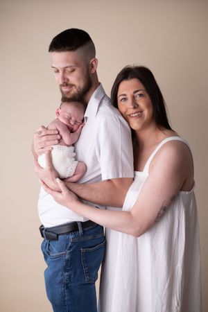 New parents with their newborn baby snuggled on dads chest with a cream background