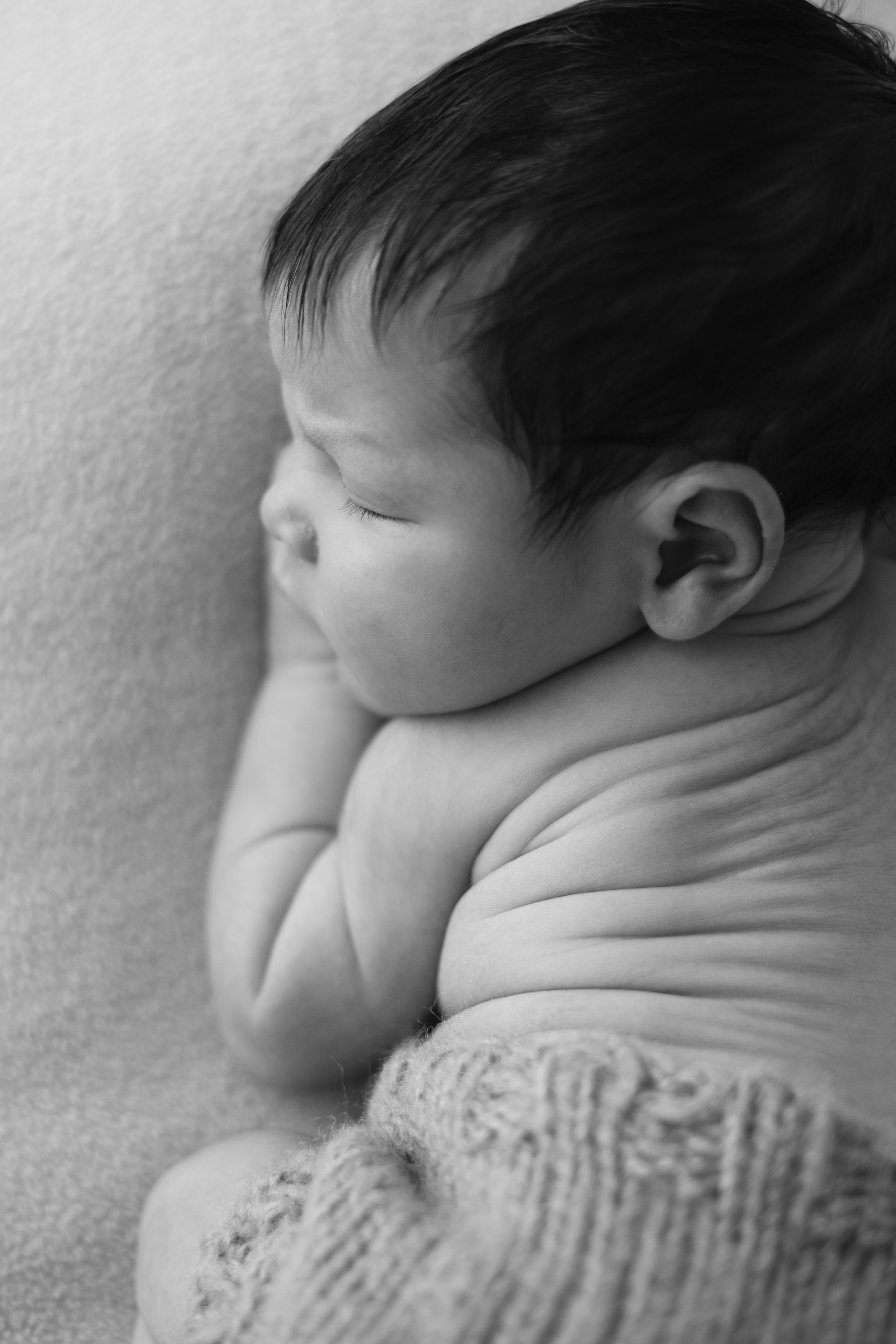 A sleeping baby wearing a knit sweater rests peacefully on a white blanket in a black and white photograph.