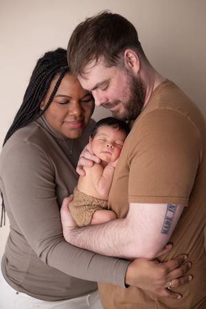 A tender family portrait shows parents embracing their newborn baby in warm brown tones against a neutral background.