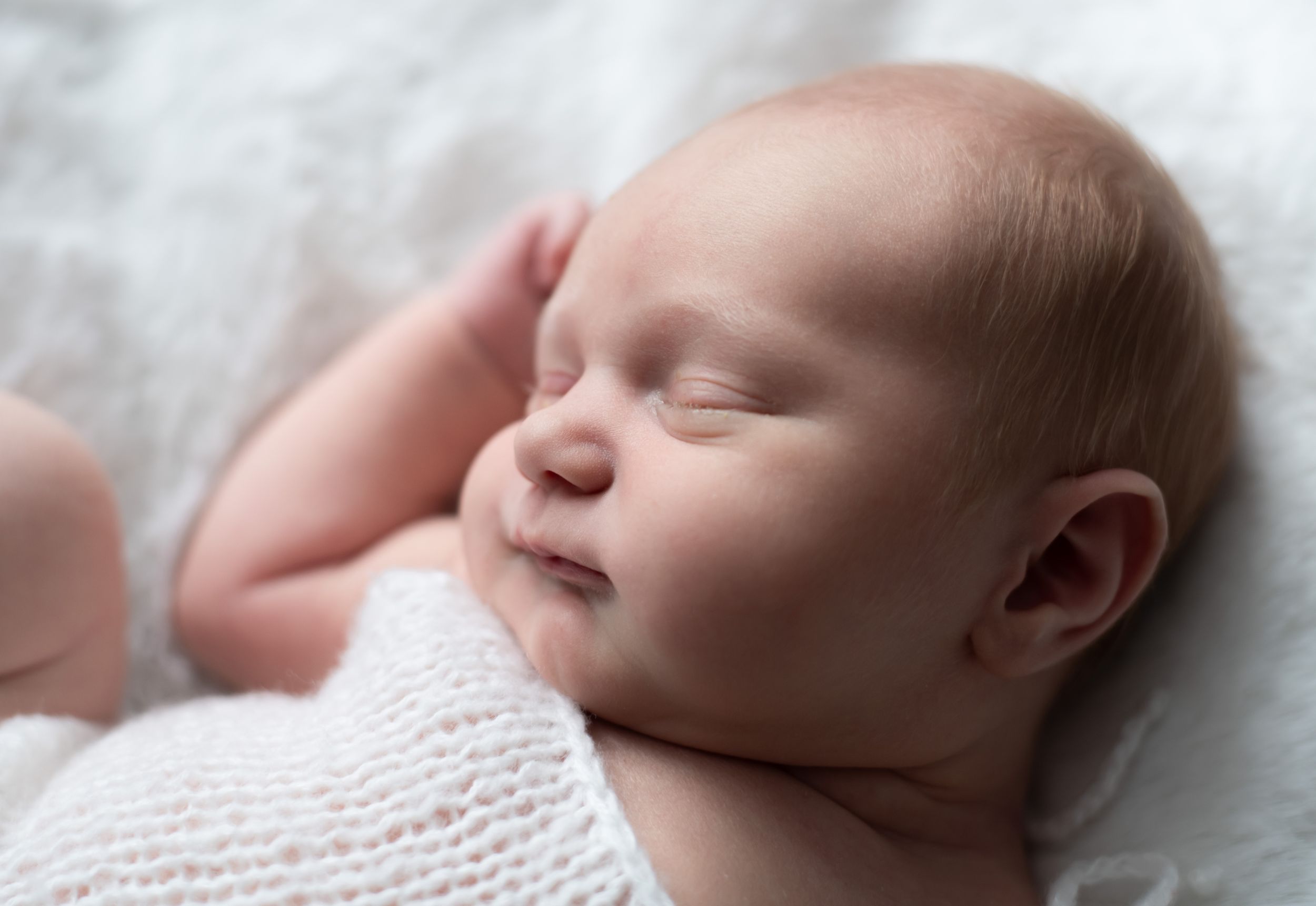 A peaceful sleeping newborn wrapped in soft white knit blanket resting on light bedding.