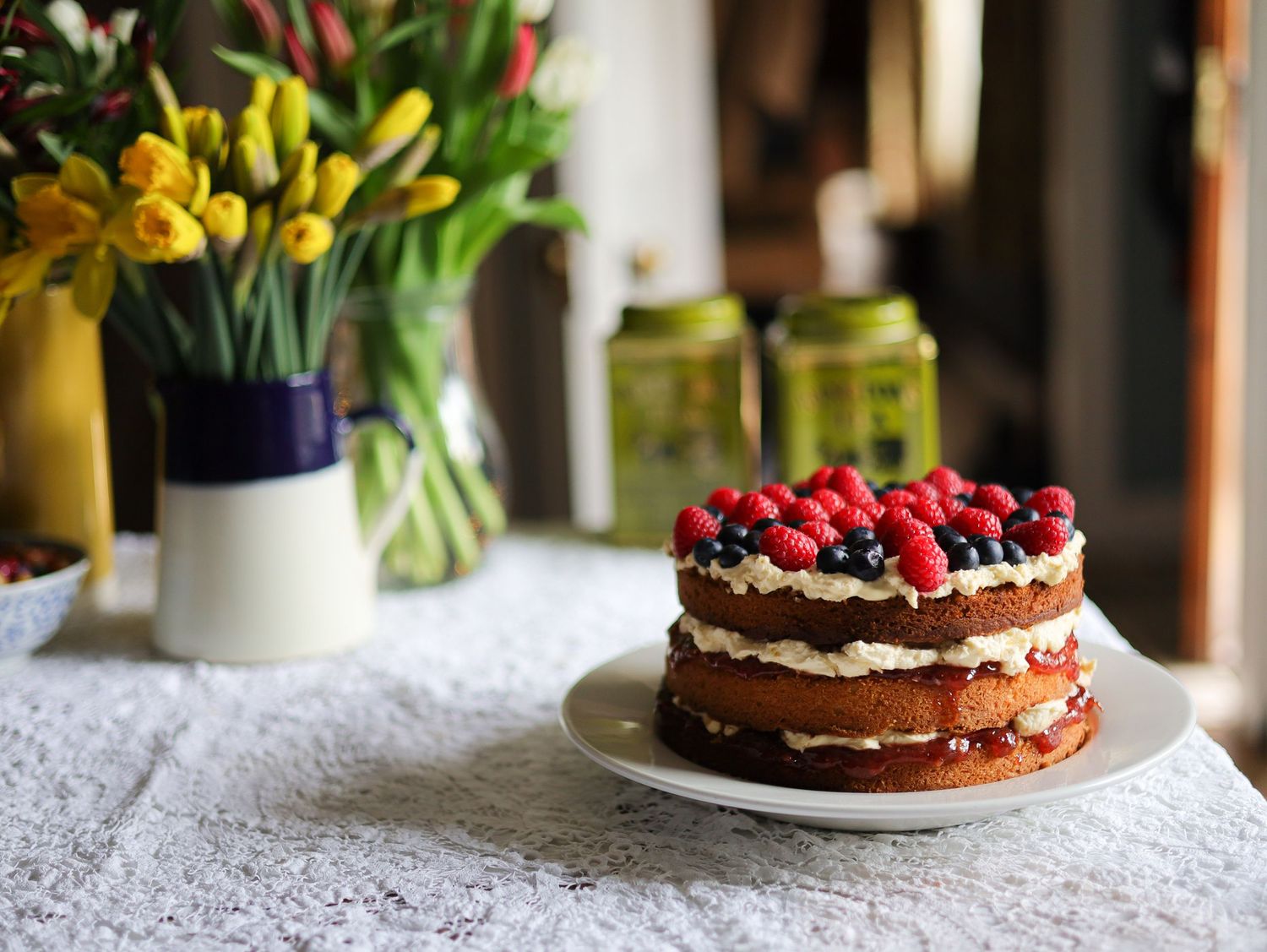 A chocolate layer cake decorated with fresh berries and flowers on a white cake stand.