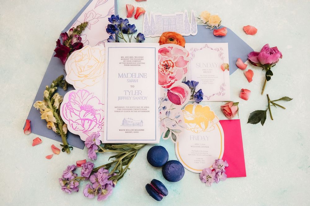 Elegant pink wedding invitations styled with colorful flowers, ribbons and macarons on a light marble surface.
