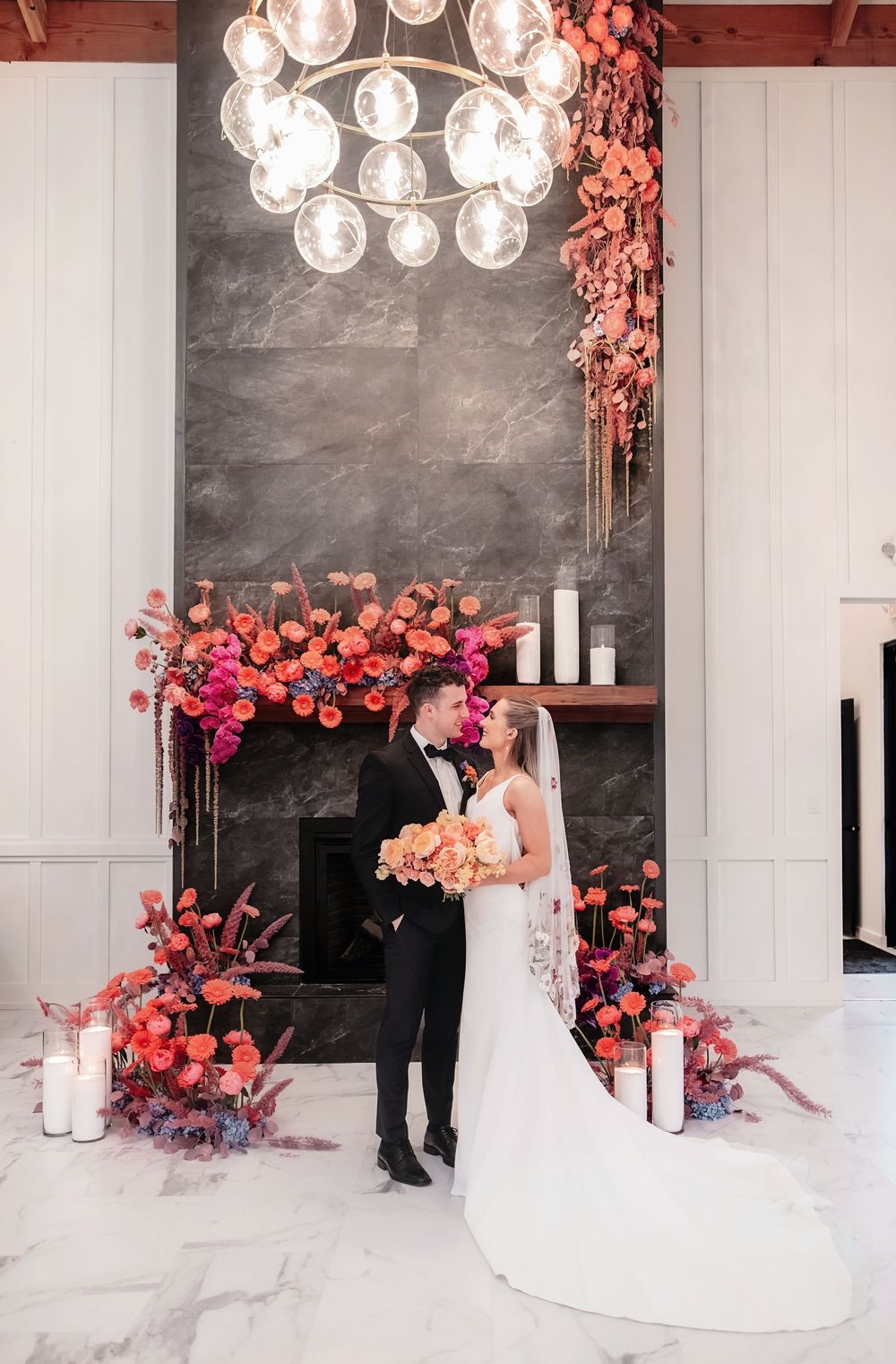 A wedding couple stands in front of a modern fireplace decorated with pink and coral floral arrangements and candles. White Willow Meadows