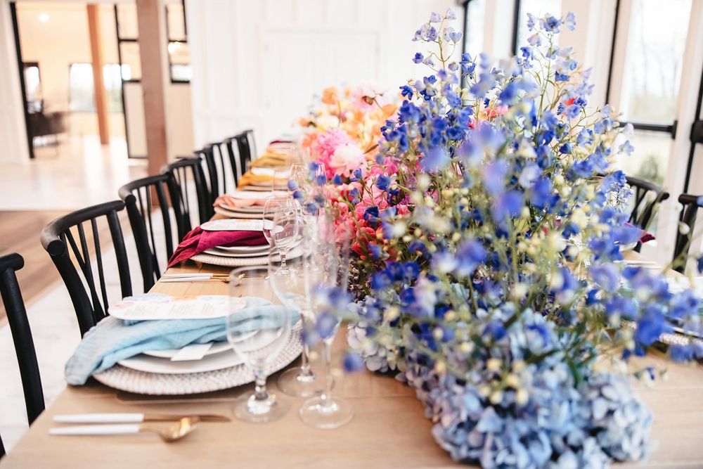 Long dining table decorated with vibrant blue and purple wildflowers in a bright, window-lit space.