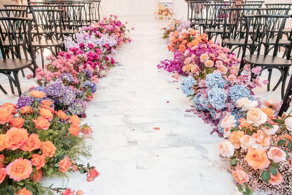Colorful floral aisle decorations line both sides of a white runner at an indoor wedding ceremony venue.