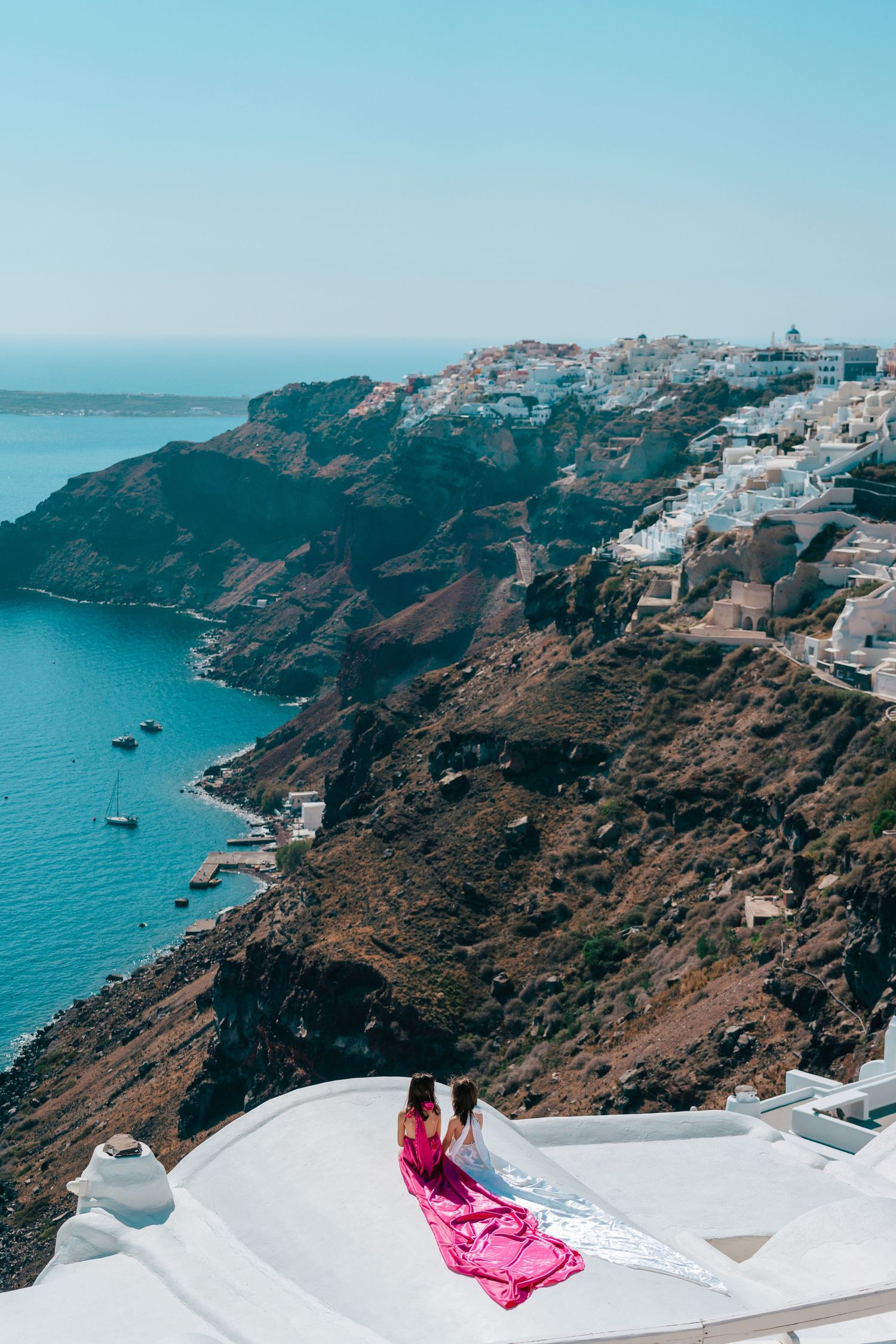 Pink flowing dress on white rooftop overlooks the dramatic coastal cliffs of Santorini.