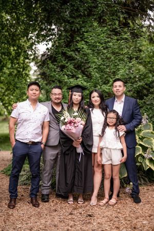 A group sits together on brown mulch ground against green foliage backdrop during graduation celebration.