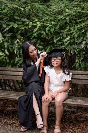 Two figures sit on a wooden bench under lush green foliage during a graduation photo session.