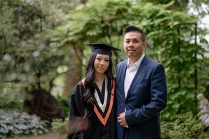 A graduation photoshoot in a lush green outdoor setting shows people wearing black cap and gown with orange accents.