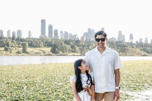 A man and young girl pose smiling by a lake with a city skyline backdrop on a sunny day.