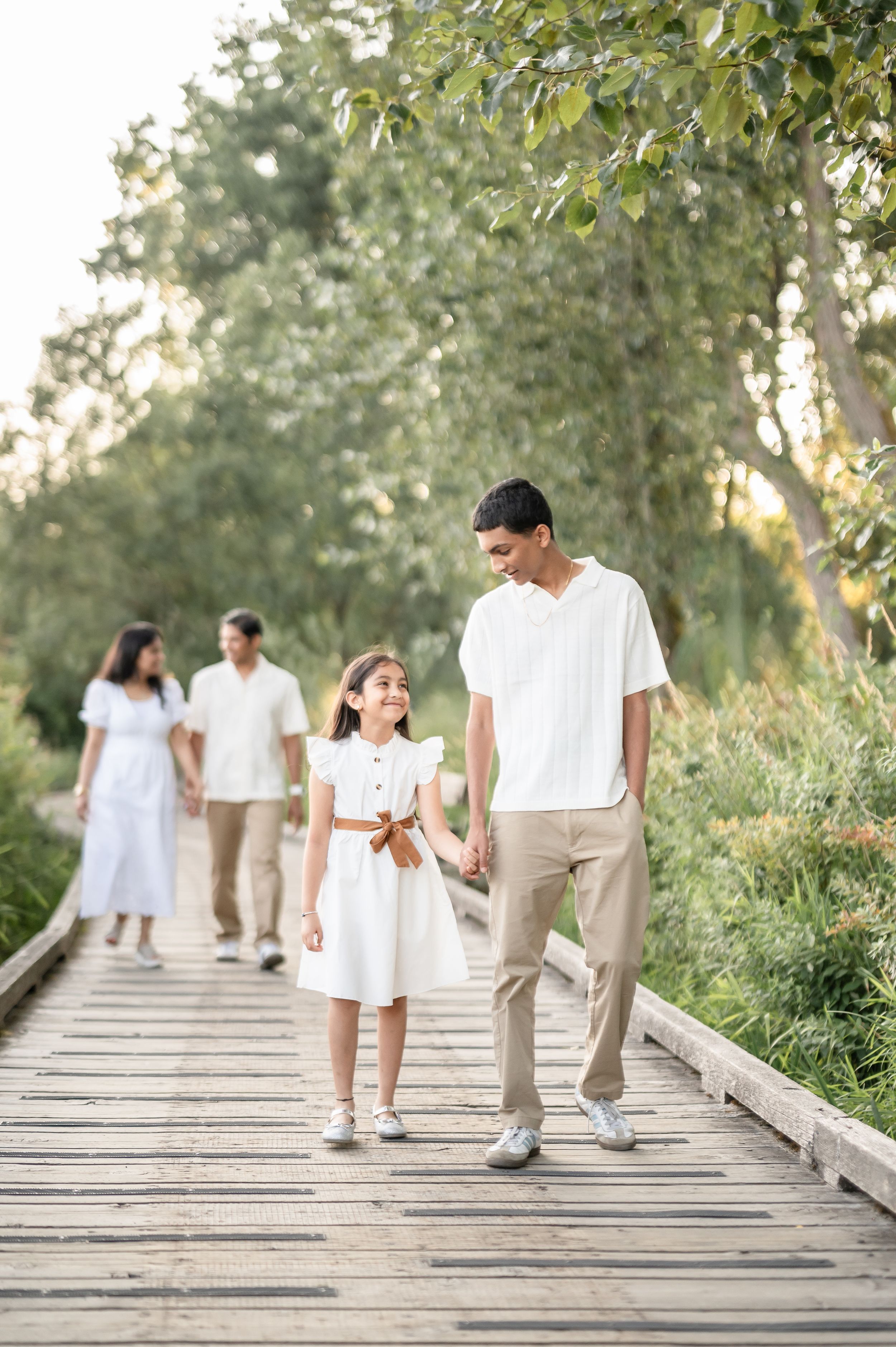 Family in white outfits walking on a wooden boardwalk path surrounded by lush green trees in a park.