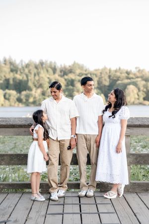 Family of four in white outfits posing on a wooden deck near a lake surrounded by lush green trees.