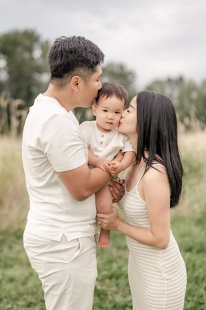 A couple in white outfits kisses their baby on the cheeks outdoors in a lush green field.