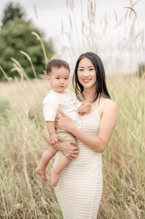 A smiling Asian woman in a white dress holds her baby boy in a sunlit field of tall golden grass.