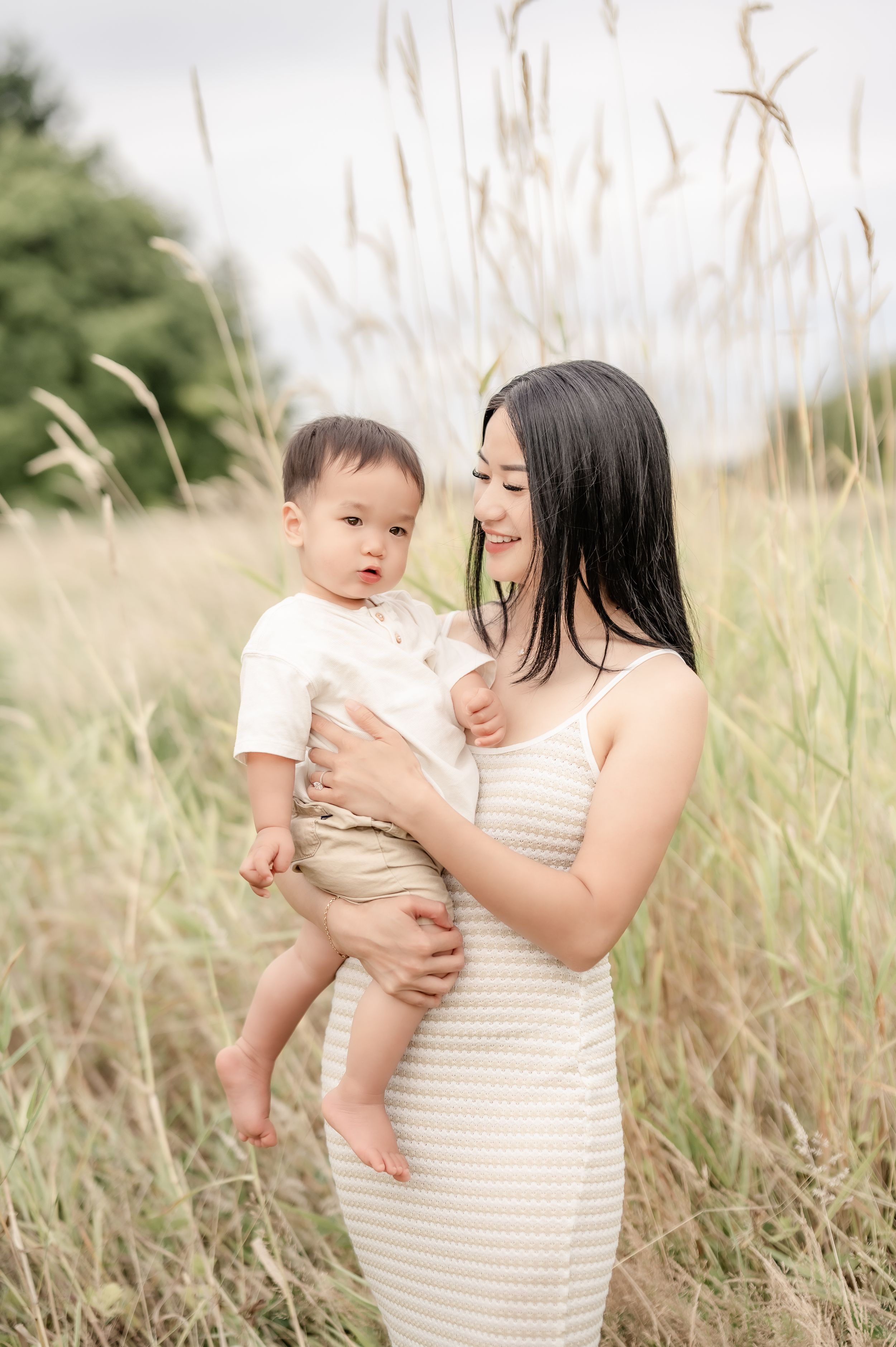 A young Asian mother in a beige dress holds her baby in a sunlit field of tall golden grass.
