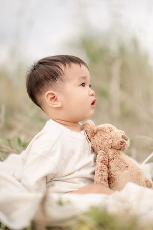 A cute Asian baby in a white outfit sits on a blanket outdoors, holding a brown teddy bear.