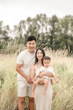 Young Asian family of three poses in tall golden grass field, parents smiling while mother holds baby in white outfit.