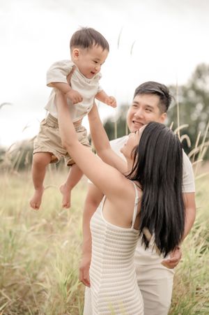 Happy Asian family playing outdoors in a field, parents lifting smiling toddler in the air on a sunny day.