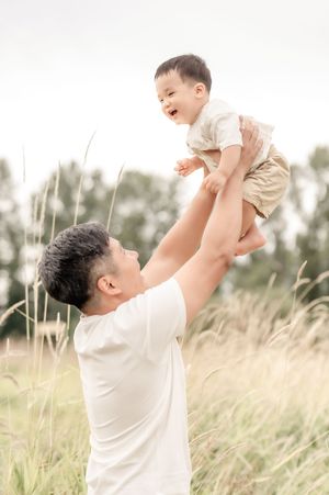 A father joyfully lifts his laughing baby boy above his head in a sunlit field with tall grass.