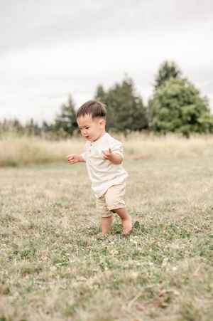 Toddler boy in beige outfit walking barefoot on green grass field with trees in background.