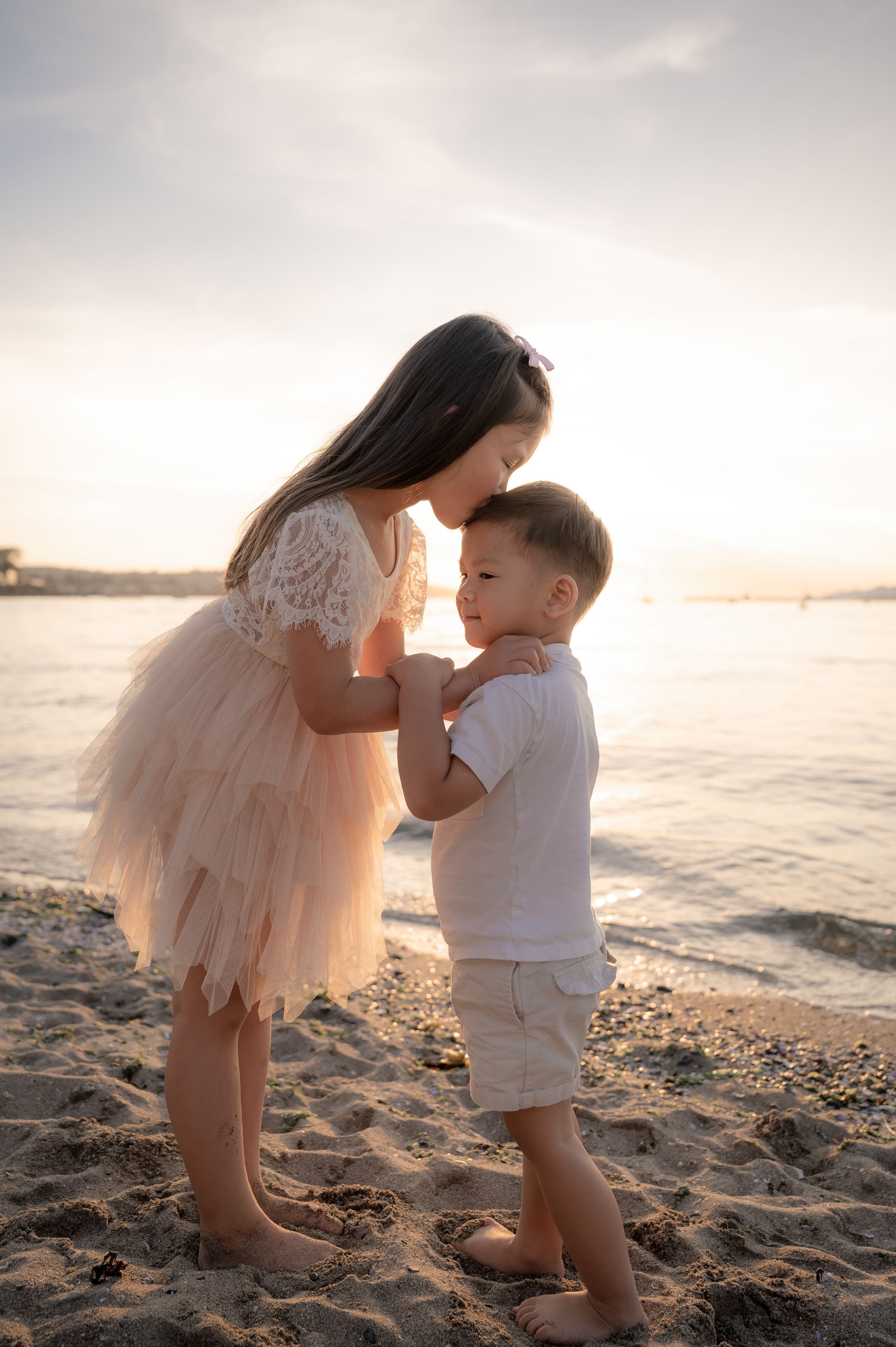 Two people share a tender moment on beach at sunset in white and light colored clothing.